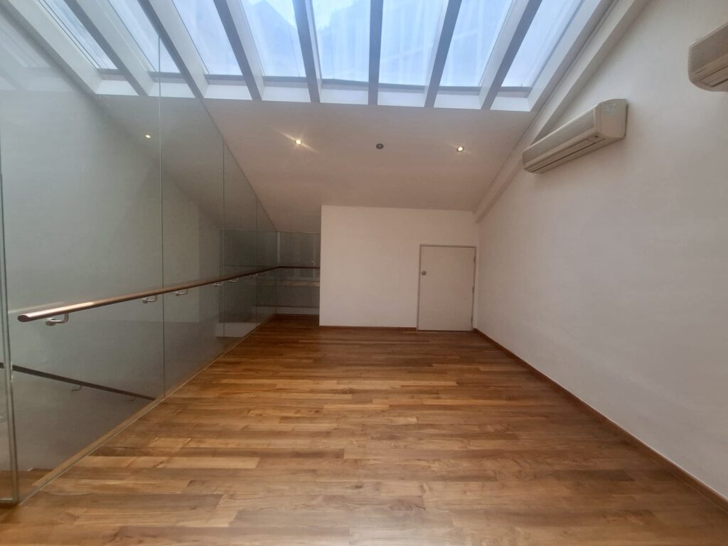 Empty room with wooden floor, glass balustrade on the left, white walls, a small door at the end, and large rooflights overhead letting in natural light.