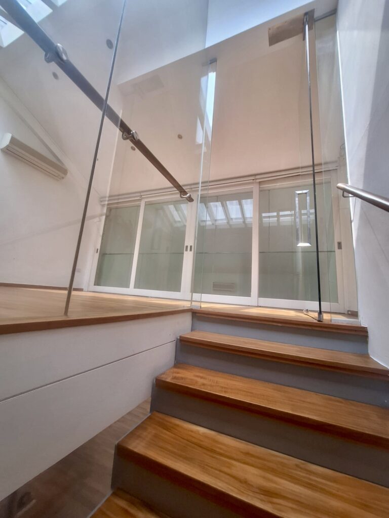 Wooden stairs with glass balustrades lead up to a landing with large windows and sliding glass doors; natural light enters from above.