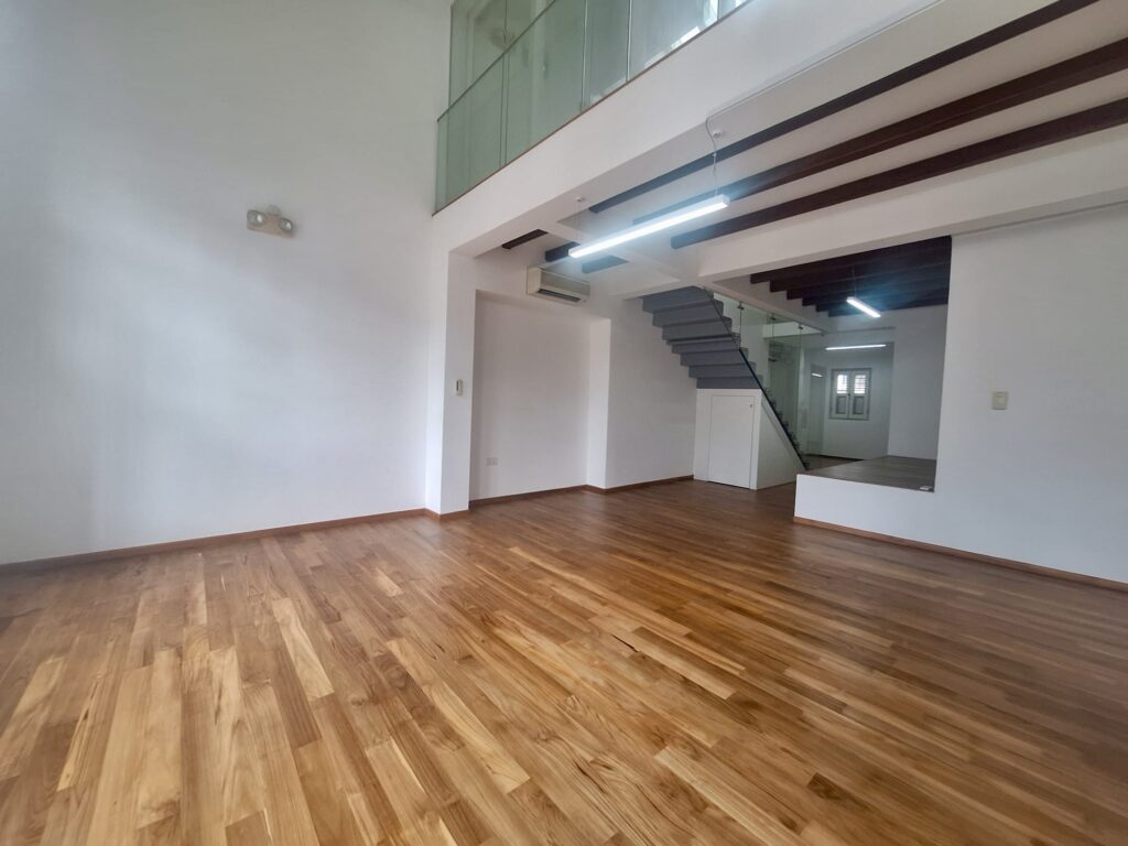 Spacious empty room with wooden flooring, white walls, exposed beams, staircase, and glass balustrade on an upper mezzanine level.