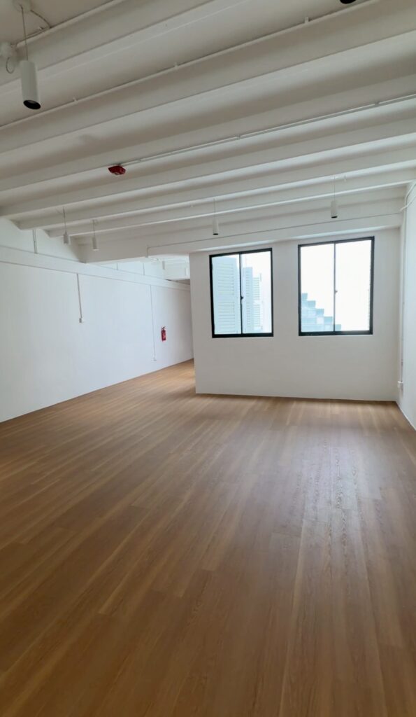 Empty room with white walls, wooden floor, two large windows letting in natural light, and an exposed beam ceiling. A red fire extinguisher is mounted near the back wall.