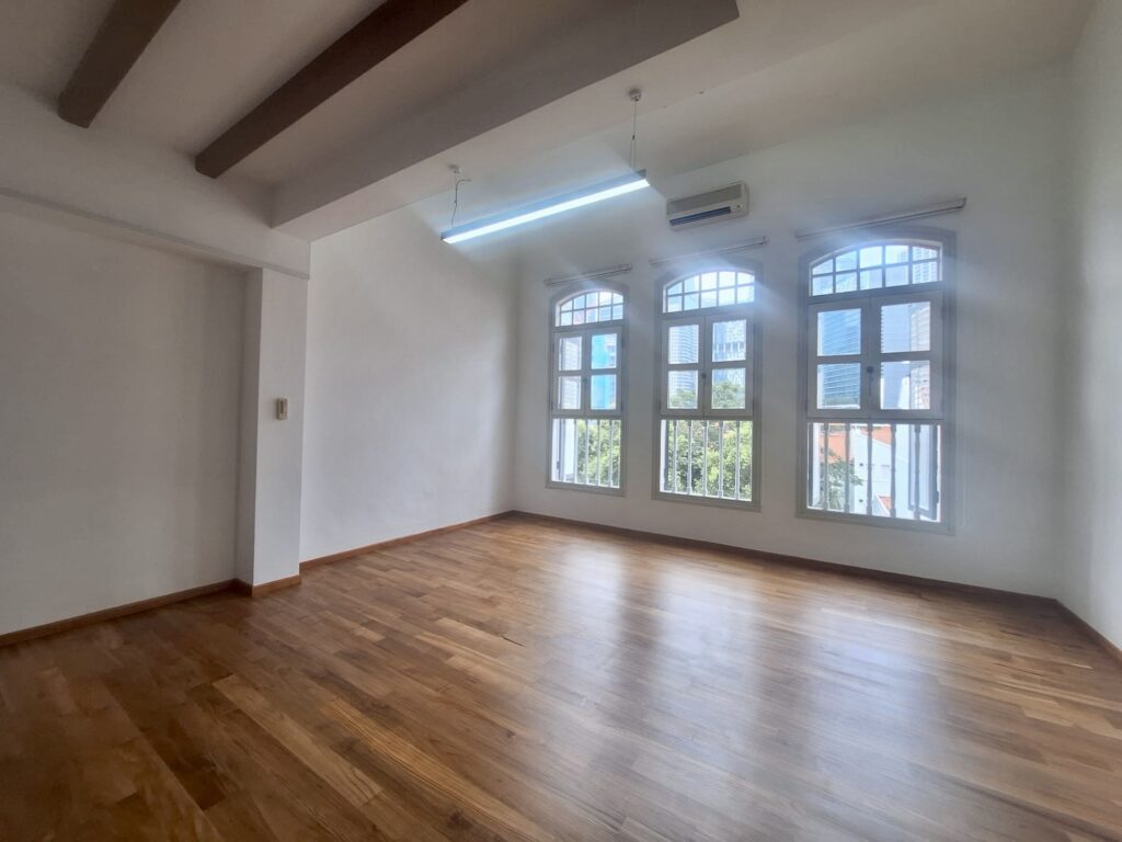Empty room with wooden flooring, white walls, exposed ceiling beams, large windows, and a wall-mounted air conditioner. Natural light enters through the windows.