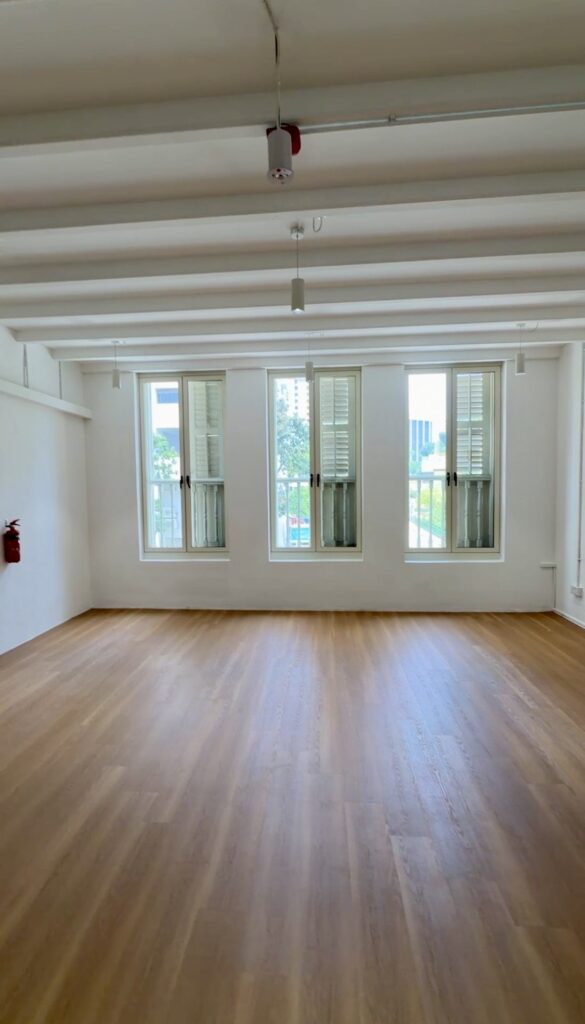 Empty room with light wood flooring, white walls, three tall windows with partially open shutters, and a fire extinguisher mounted on the left wall.