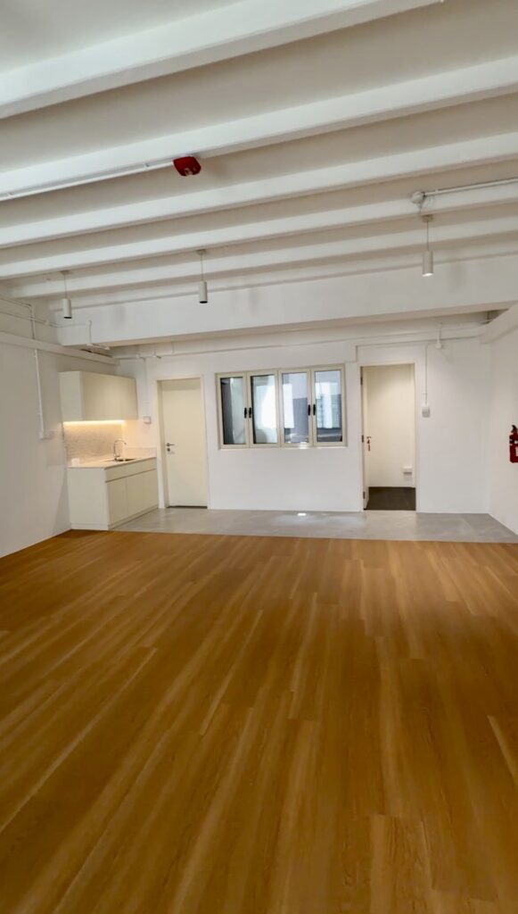 Empty room with wooden flooring, white walls, exposed ceiling beams, a kitchenette on the left, windows in the centre, and a fire extinguisher mounted on the right wall.