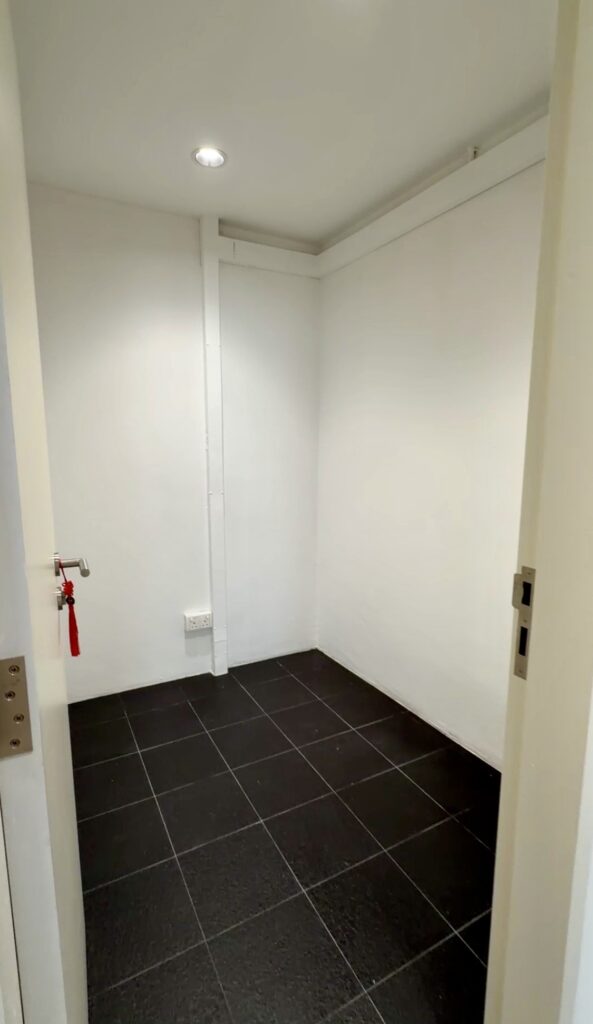 Small, empty room with white walls, black tiled floor, a ceiling light, and a closed white door partially visible on the right. A key with a red string hangs from the left door handle.