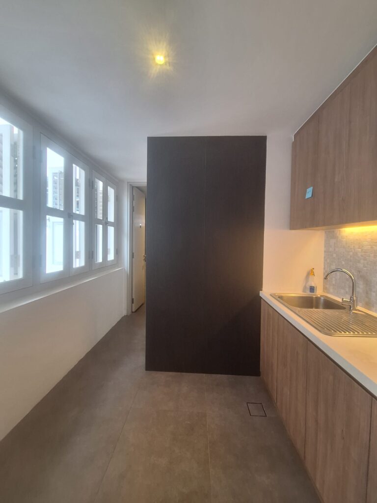 A narrow kitchen with wooden cupboards, a sink, under-cupboard lighting, large windows on the left, and a dark door at the end of the room.