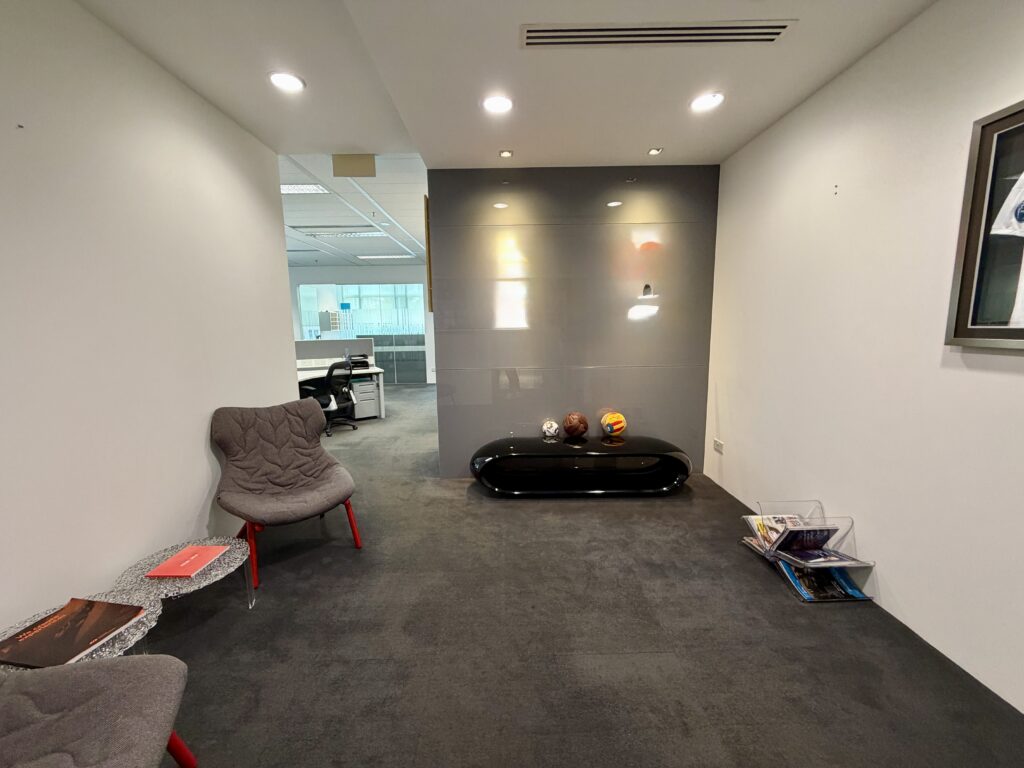 Modern office waiting area with two chairs, a side table with magazines, a glossy black console with decorative items, and a magazine rack against white and grey walls.