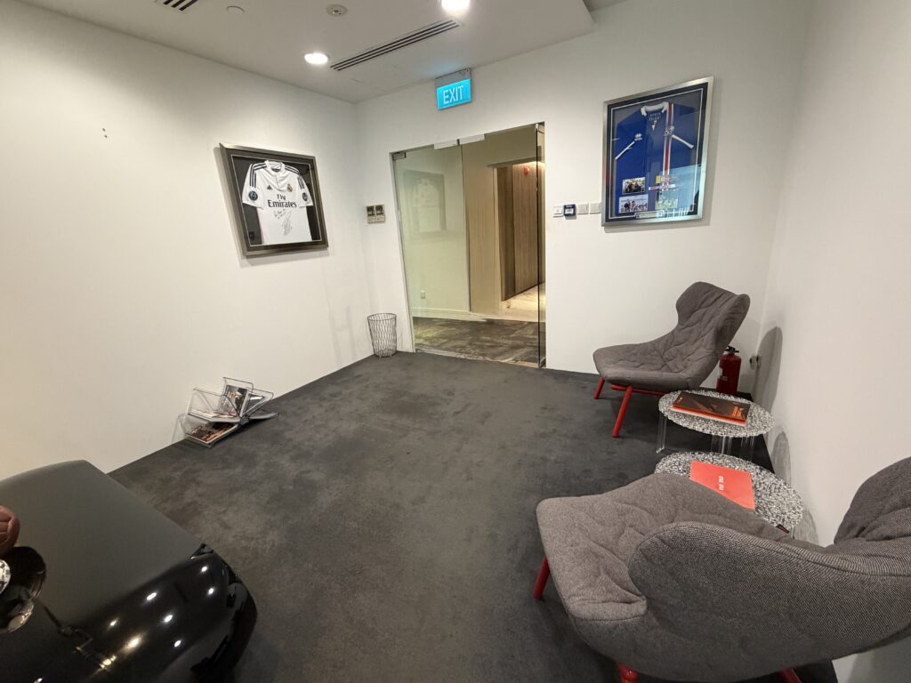 A small waiting area with two grey chairs, a black table, framed sports jerseys on the wall, a wire magazine rack, and a glass door leading to a corridor.