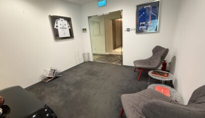 A small waiting area with two grey chairs, a black table, framed sports jerseys on the wall, a wire magazine rack, and a glass door leading to a corridor.