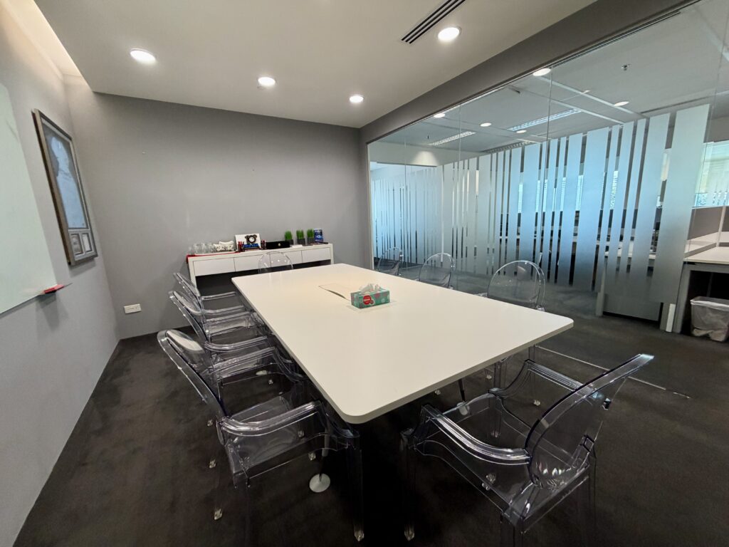 A modern conference room with a white table, six clear chairs, a whiteboard, and a glass wall. Office supplies and drinks are arranged on a side cupboard.