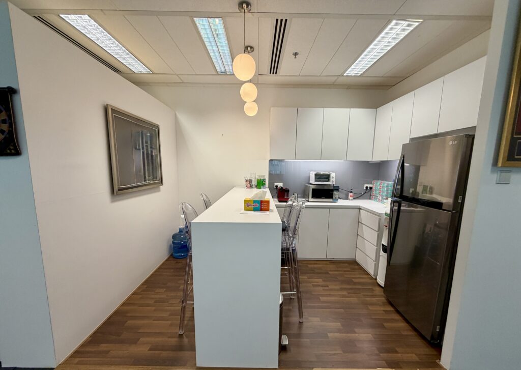 Modern office kitchenette with white cupboards, a tall table with clear chairs, a stainless steel fridge, kitchen appliances, and ceiling lights on a wooden floor.