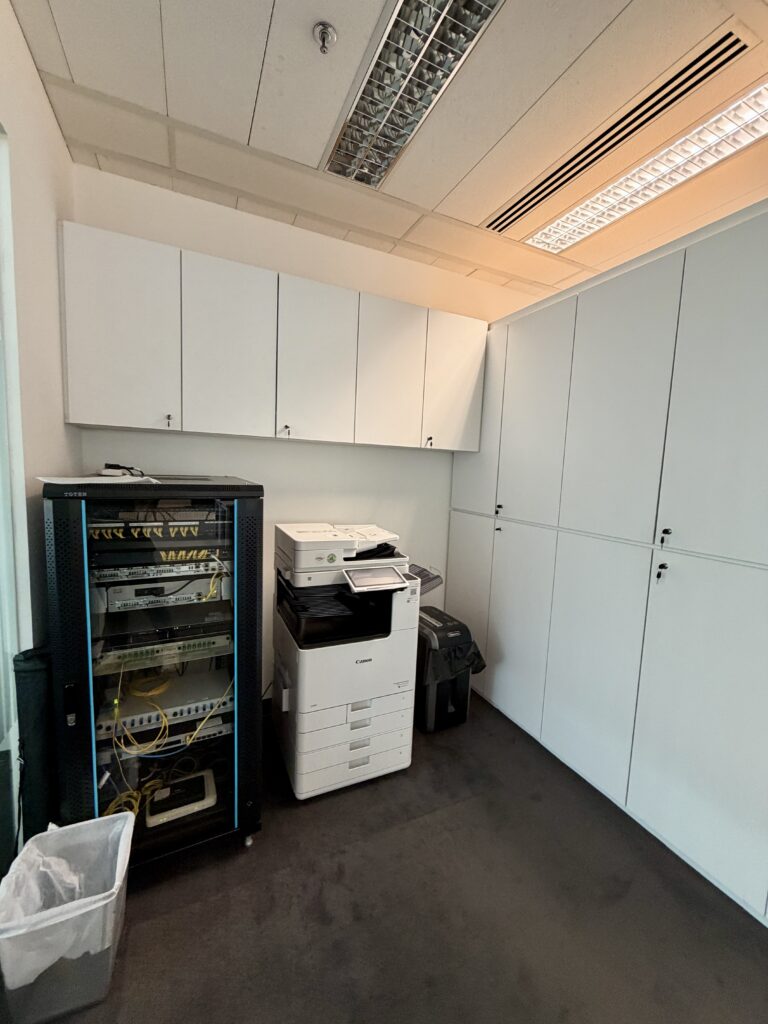 Small office room with network equipment in a rack, a photocopier/printer, a shredder, rubbish bin, and white storage cupboards along the walls.