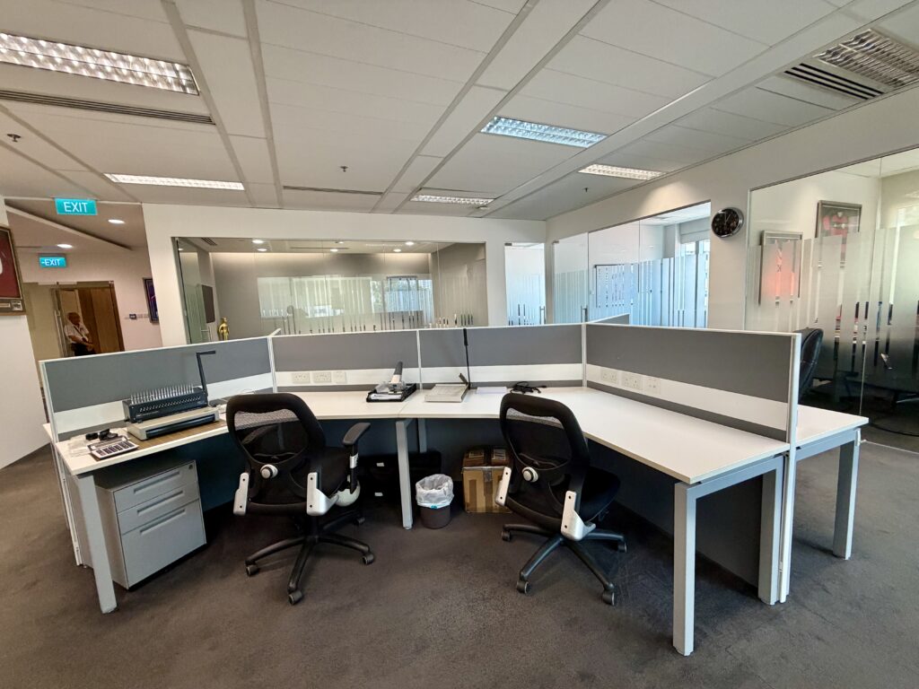 A modern office workspace with three empty desks, black chairs, office supplies, and a rubbish bin, set in a room with glass walls and a clock on the wall.