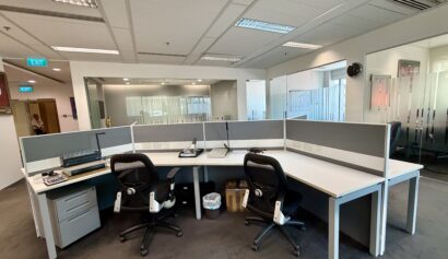 A modern office workspace with three empty desks, black chairs, office supplies, and a rubbish bin, set in a room with glass walls and a clock on the wall.