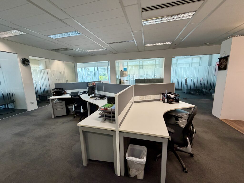 Modern office with empty workstations, computers, office chairs, and partitions; glass-walled meeting rooms in the background, and a dartboard on the wall to the right.