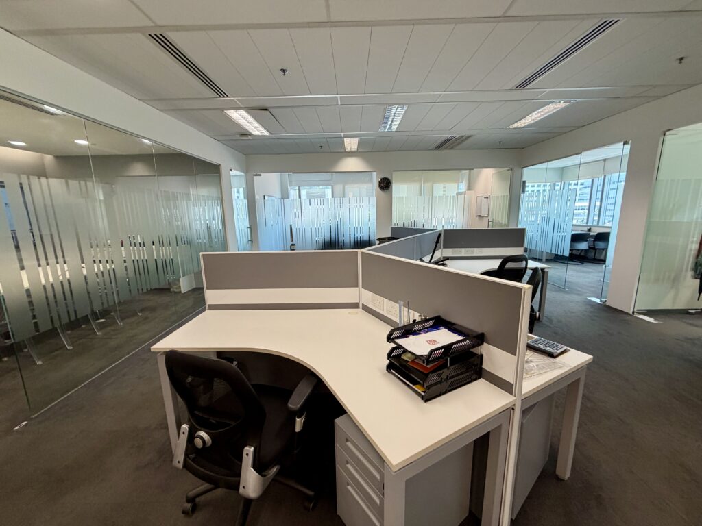 Modern office with glass walls, empty workstations, ergonomic chairs, and natural light from large windows.