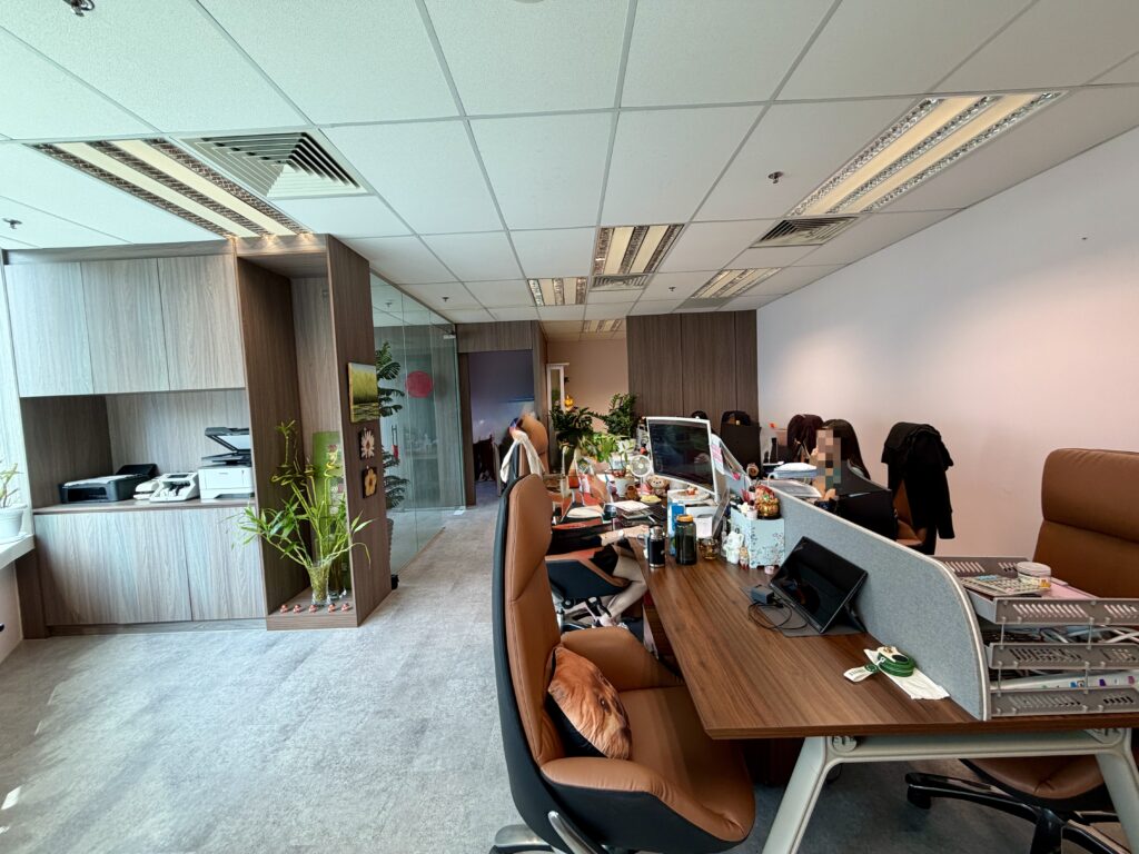 An office with rows of wooden desks and brown office chairs, separated by grey partitions.