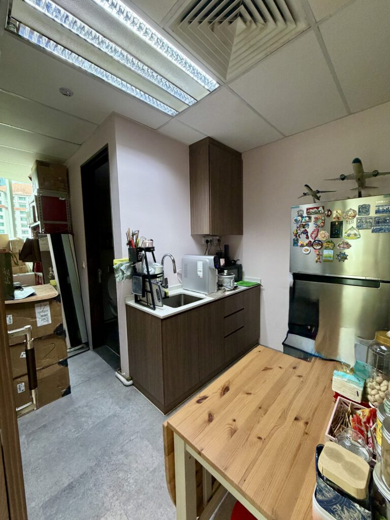 A small office kitchen with dark wood cupboards, a stainless steel sink, a fridge covered in magnets, and a wooden table in the foreground.