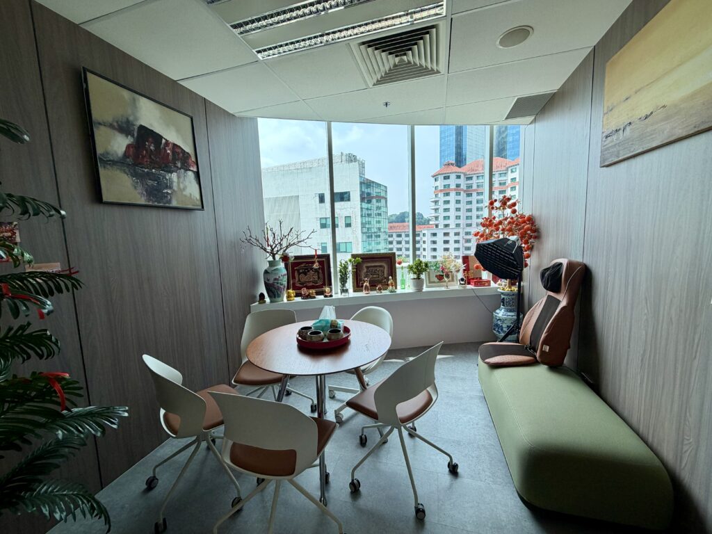A small meeting room with a round table, four chairs, and a long green bench with a massage seat cushion, overlooking a city skyline.