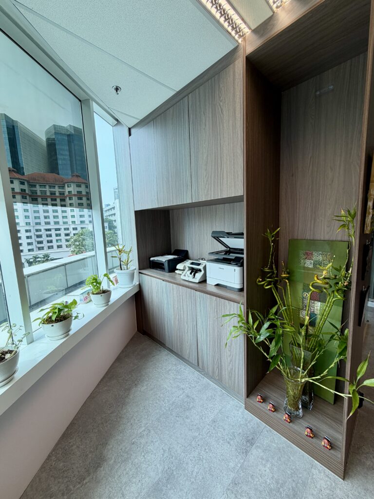 Office corner with wood-grain cupboards, a printer and scanner on a shelf, a window sill with small potted plants, and a large vase of bamboo stalks.