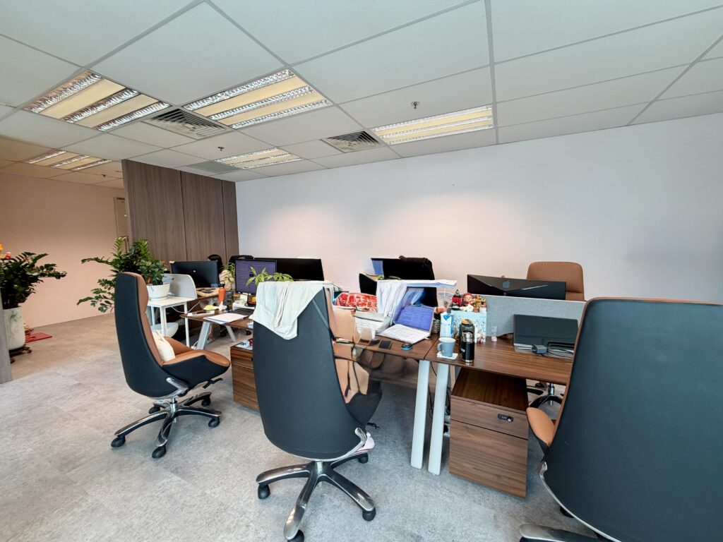 An open-plan office workspace featuring multiple desks, office chairs, computers, and personal belongings.
