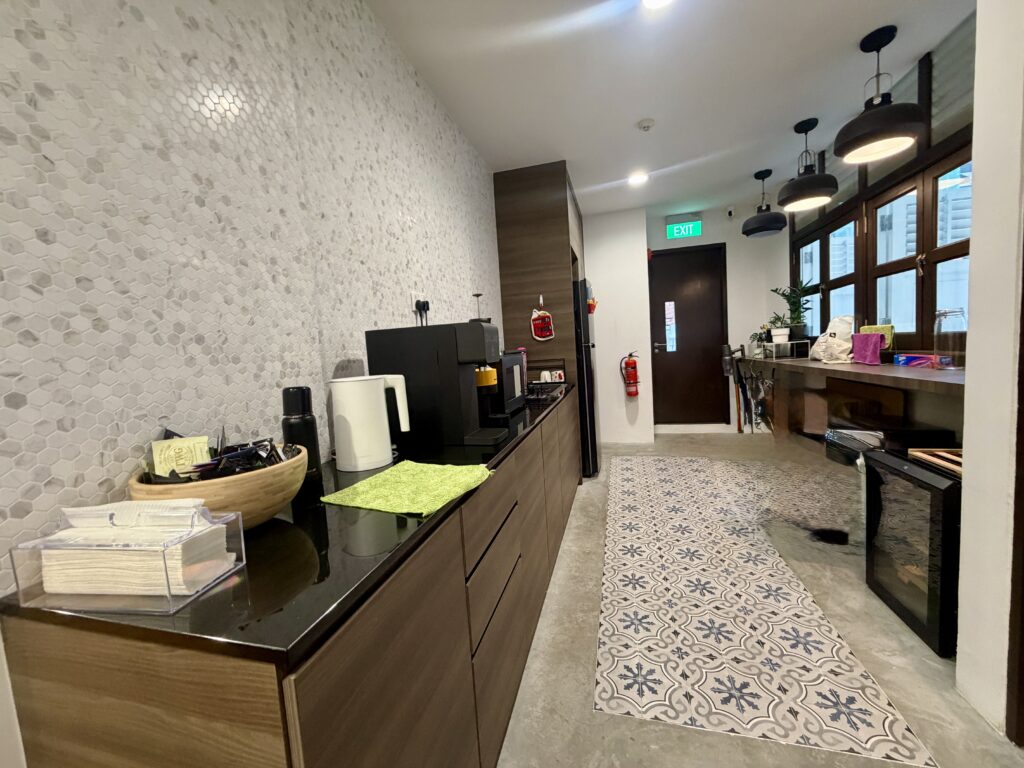 A narrow office kitchenette with a coffee machine, a wooden bowl of tea bags, and a patterned runner rug on the floor.