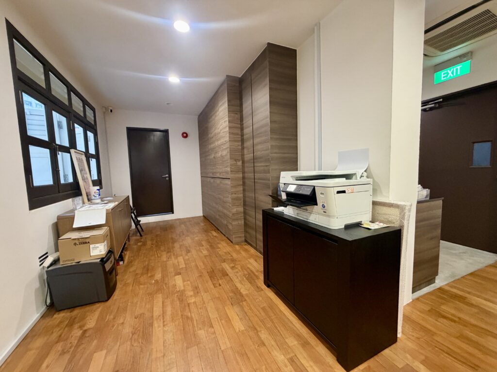 Office corridor with wooden flooring, a wooden storage unit, a printer on a black cupboard, and an exit sign above a doorway.