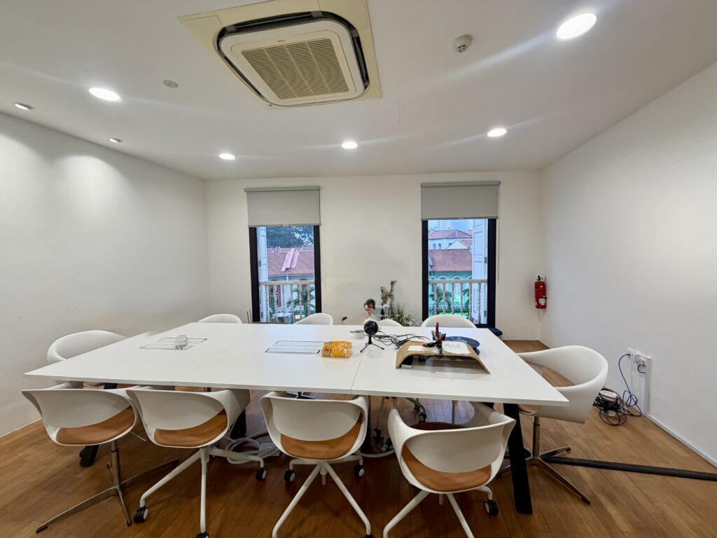 A white conference table with eight chairs in a room with two windows and a ceiling-mounted air conditioning unit.