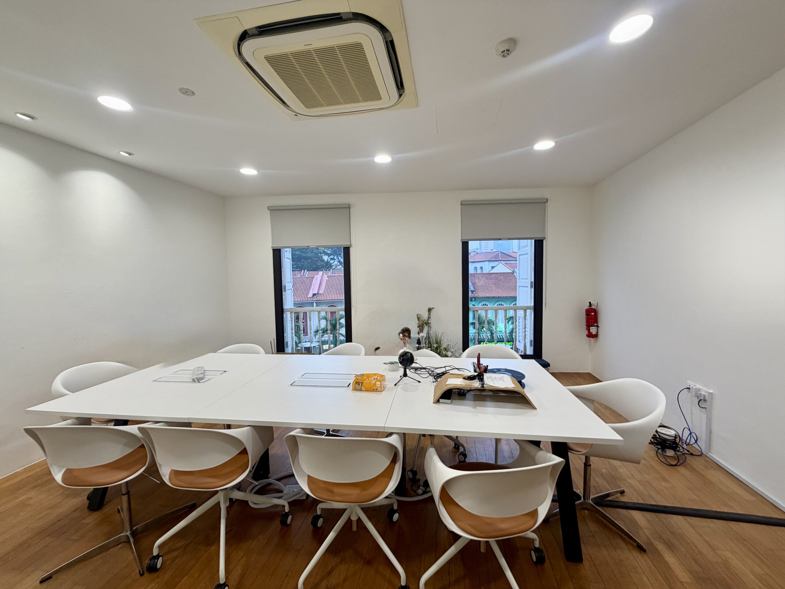 A white conference table with eight chairs in a room with two windows and a ceiling-mounted air conditioning unit.