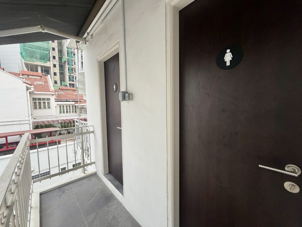 Two dark doors with circular icons indicating toilets along an outdoor corridor, overlooking city buildings.