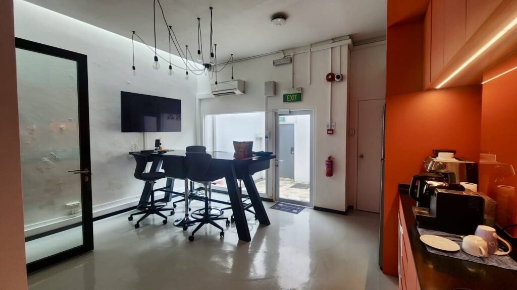 A modern office kitchen featuring a high central table with bar stools, a wall-mounted TV, and a coffee station on an orange feature wall.