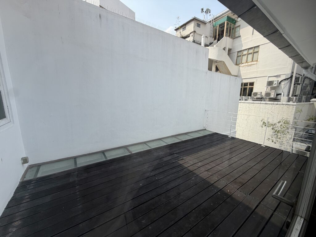 A wooden decking with a glass-panelled edge next to a tall white wall, overlooking surrounding buildings.