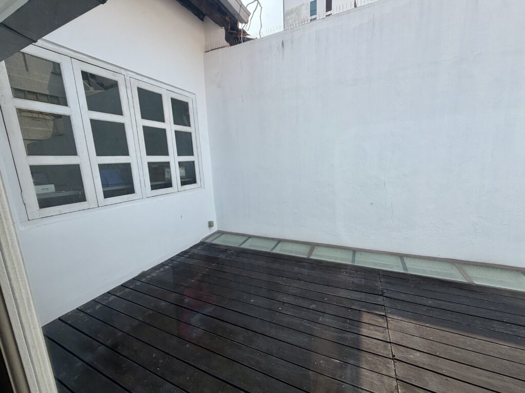 A rooftop terrace with dark wood decking, a white wall, and a window with four panes.