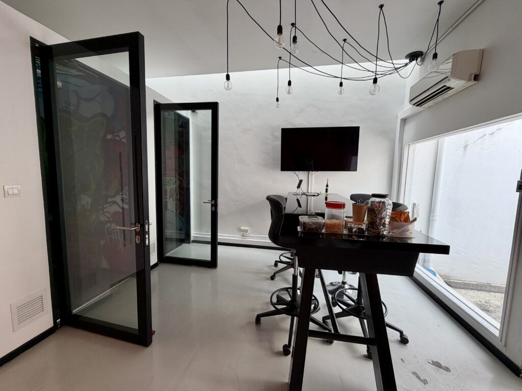 A modern office meeting room with a black high-top table, stools, a wall-mounted television, and two open black-framed glass doors.