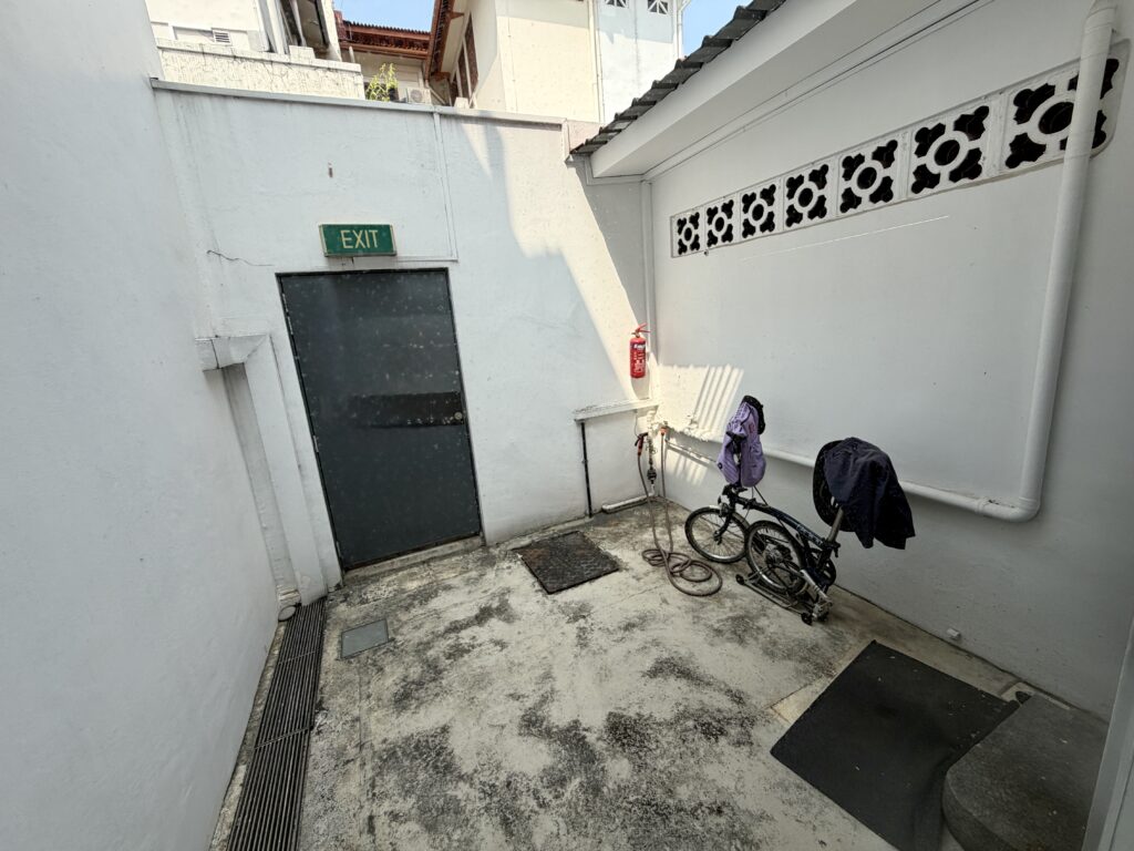 A paved alleyway with a dark door marked with a green “EXIT” sign, a fire extinguisher on the wall, and a folded bicycle with items draped over it.