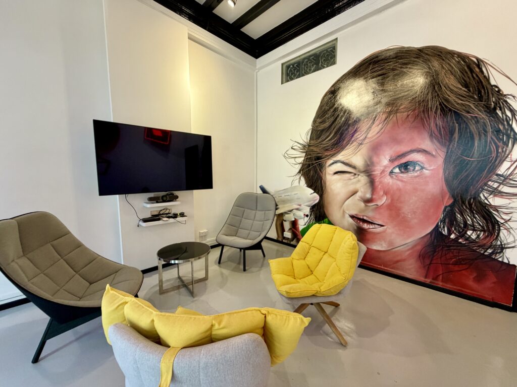 A lounge area with four modern chairs, a small round table, a wall-mounted television, and a large mural of a girl’s face.