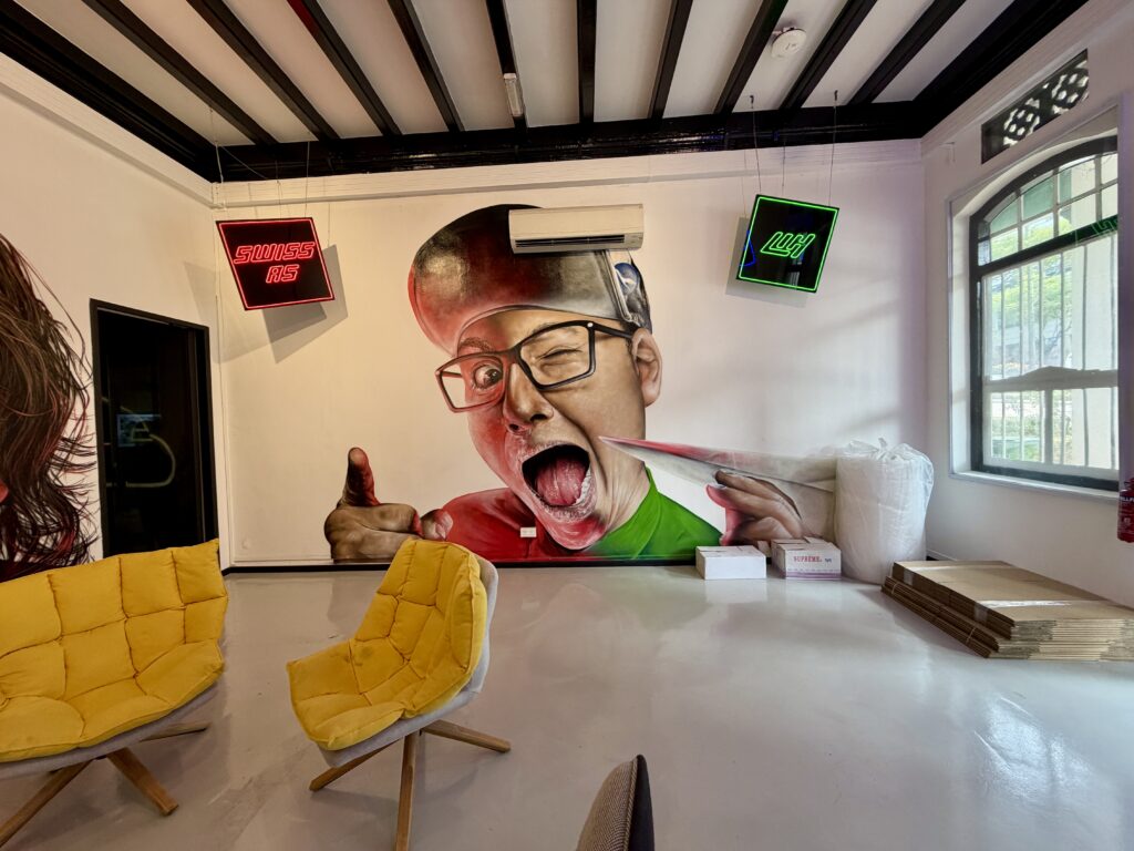 A large mural of a winking man with his mouth open and pointing, flanked by “SWISS AS” and “LGH” neon signs, above two yellow chairs.