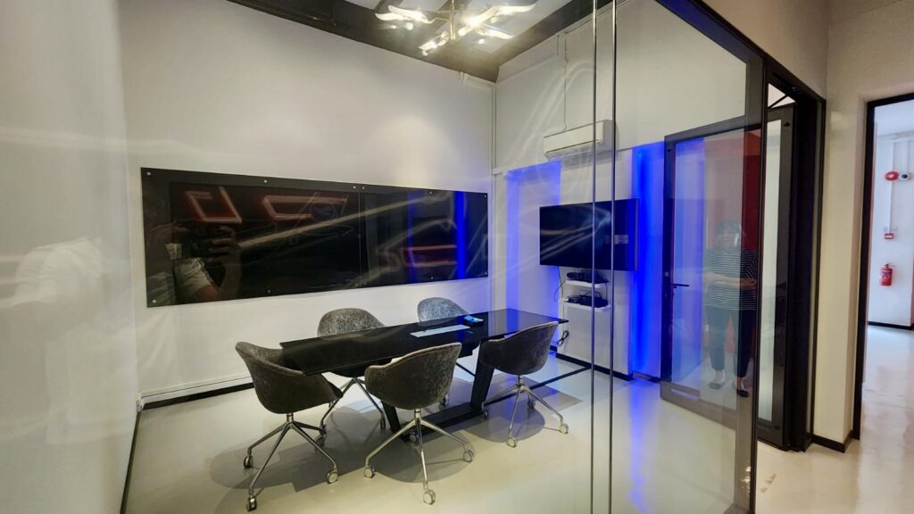 A glass-walled office meeting room featuring a black table, four grey chairs, a wall-mounted television, and a black glass whiteboard.
