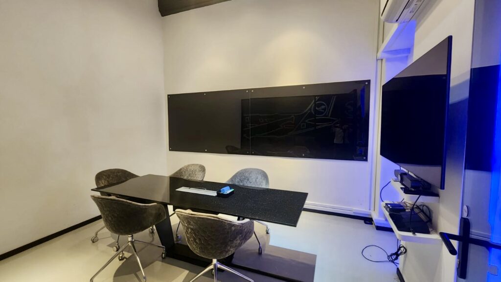 A conference room with a black rectangular table, four grey chairs, a mounted black whiteboard, and a wall-mounted television with blue accent lighting.
