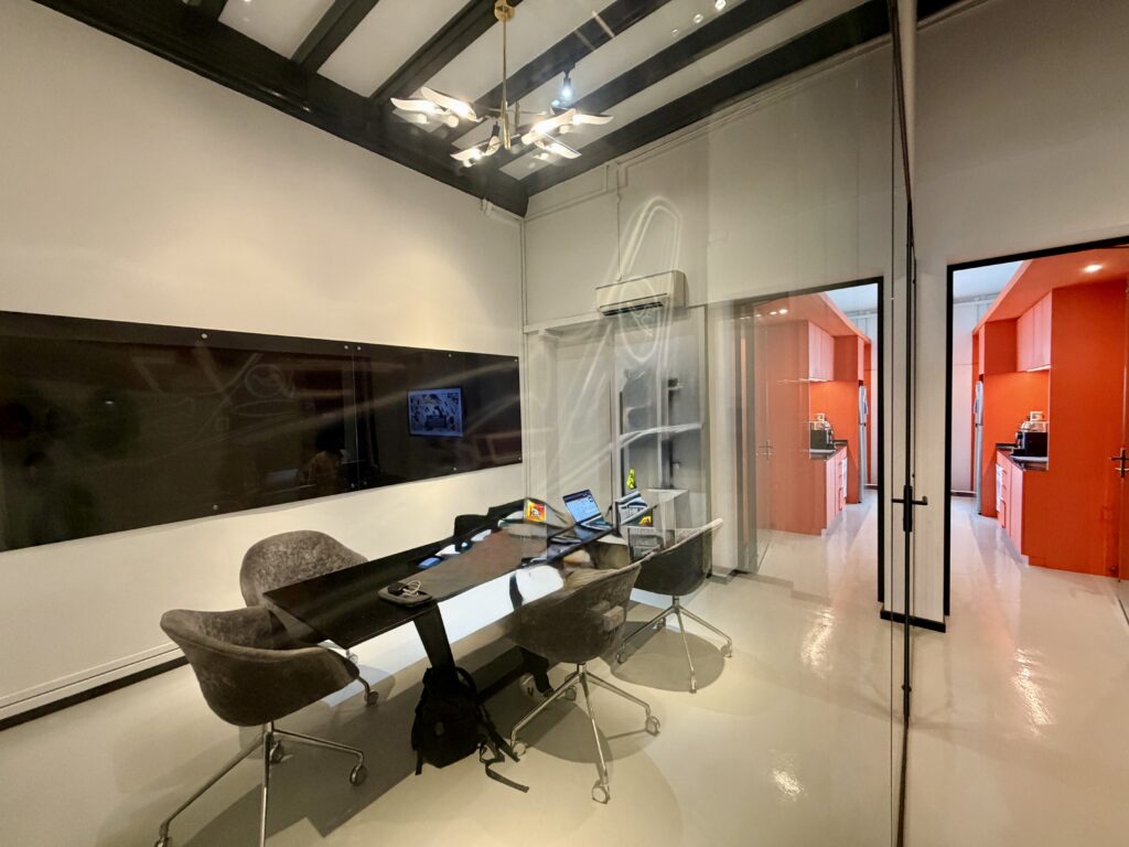 A conference room with a black table and four chairs, looking into an adjacent office with orange cupboards and walls.