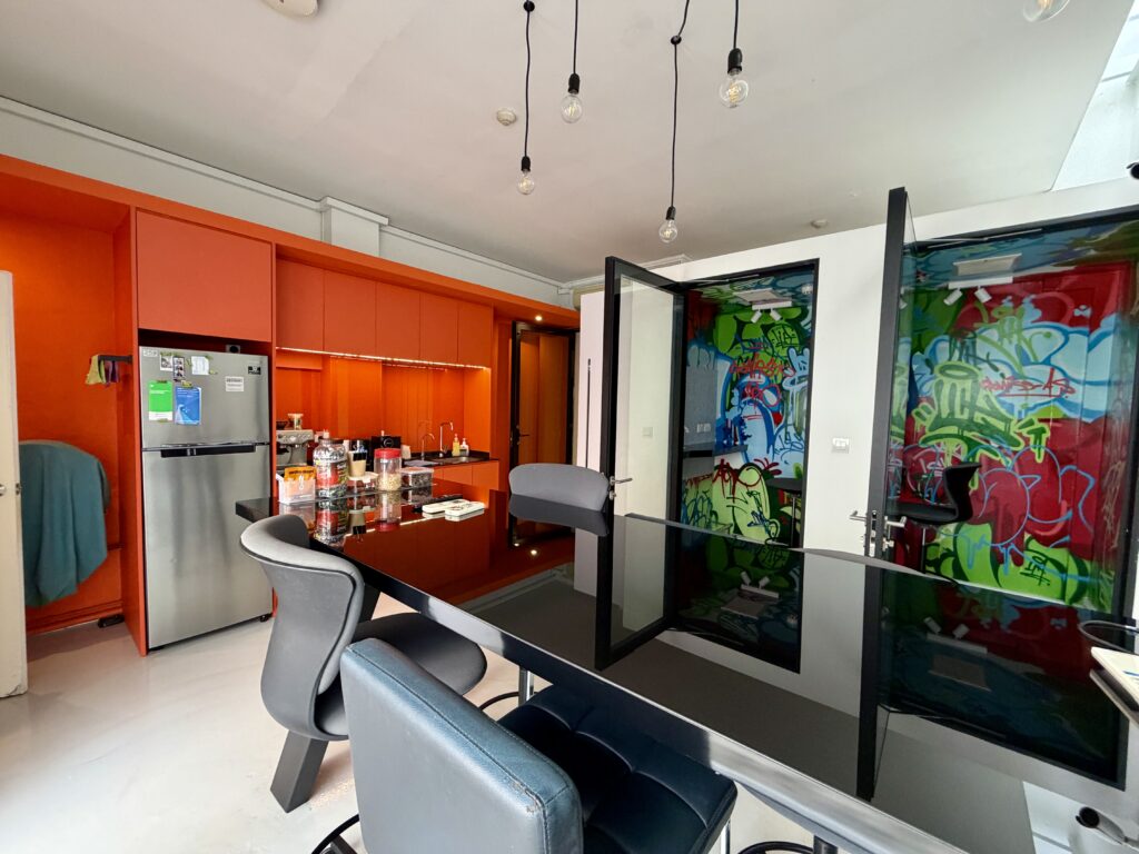 A modern kitchen with bright orange cabinetry, a stainless steel fridge, and a black dining table, featuring glass doors decorated with colourful graffiti-style art.