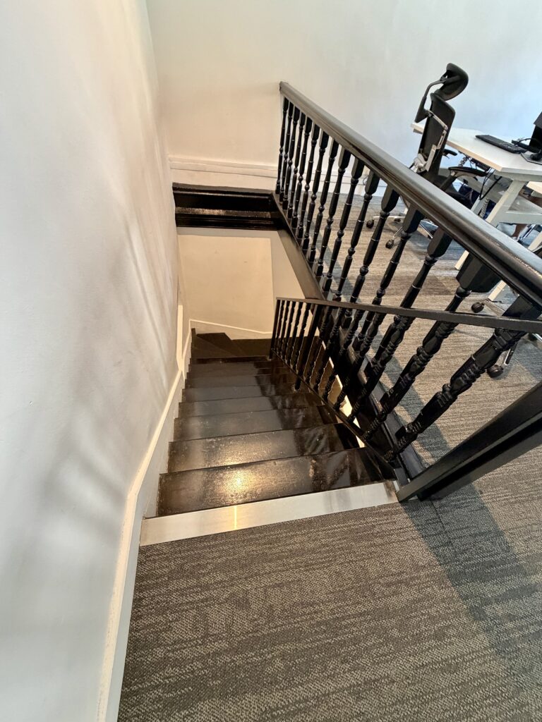 A wooden staircase with a black railing, viewed from the top landing next to an office desk.