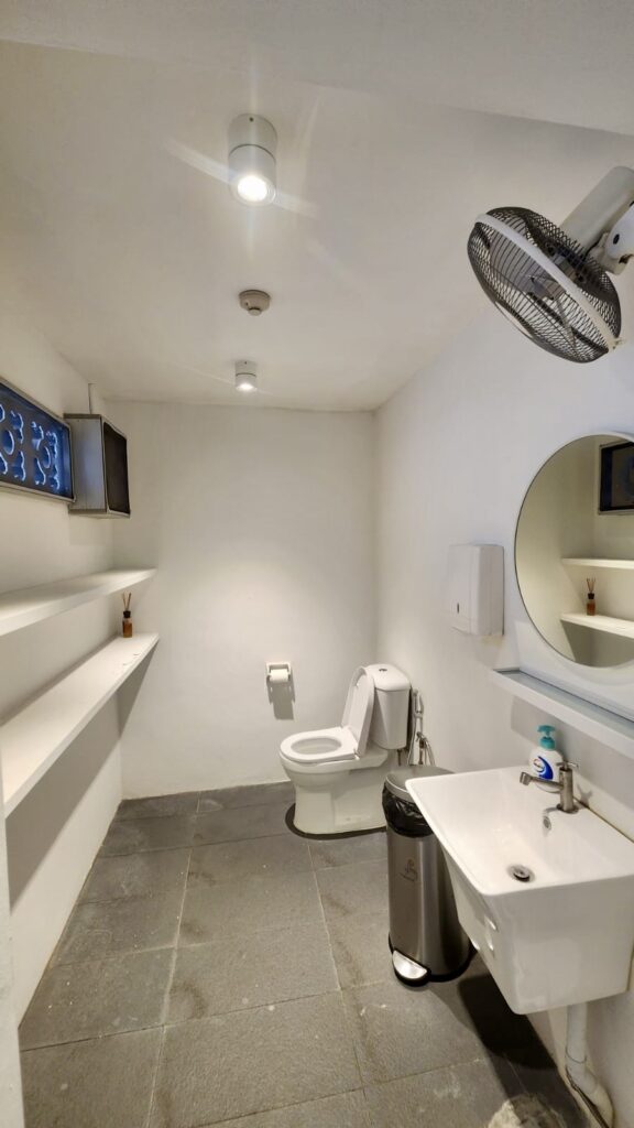 A small bathroom with white walls and grey tiled floors, featuring a toilet, a pedestal basin with a soap dispenser, a round mirror, a wall-mounted fan, and two long shelves.