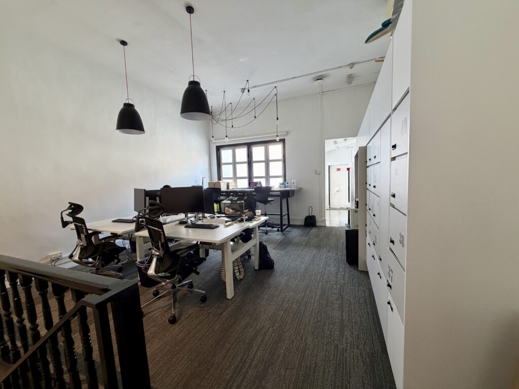 An office space with two desks, computer monitors, black office chairs, pendant lighting, and a row of numbered white storage lockers.