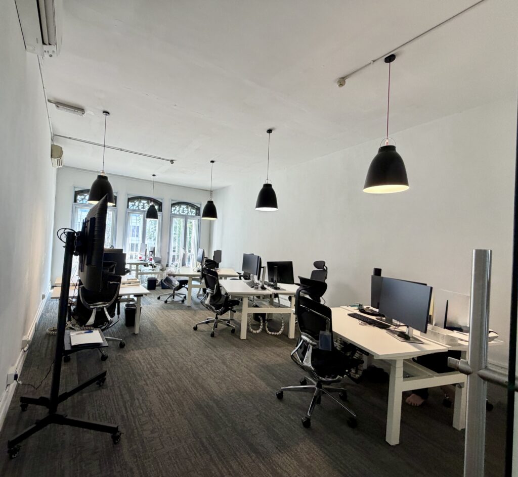 An office with rows of white desks, black office chairs, computer monitors, and hanging pendant lights.