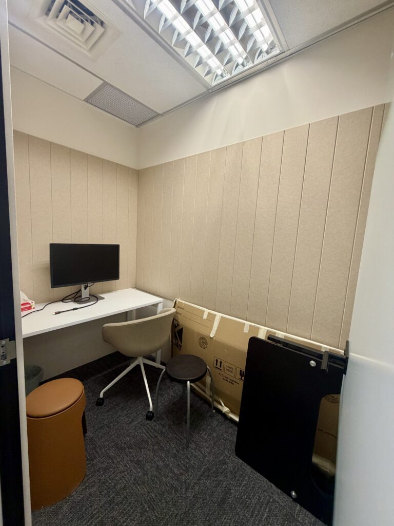 Small office space with a desk, computer monitor, wheeled office chair, two small stools, and a large cardboard box leaning against the wall.