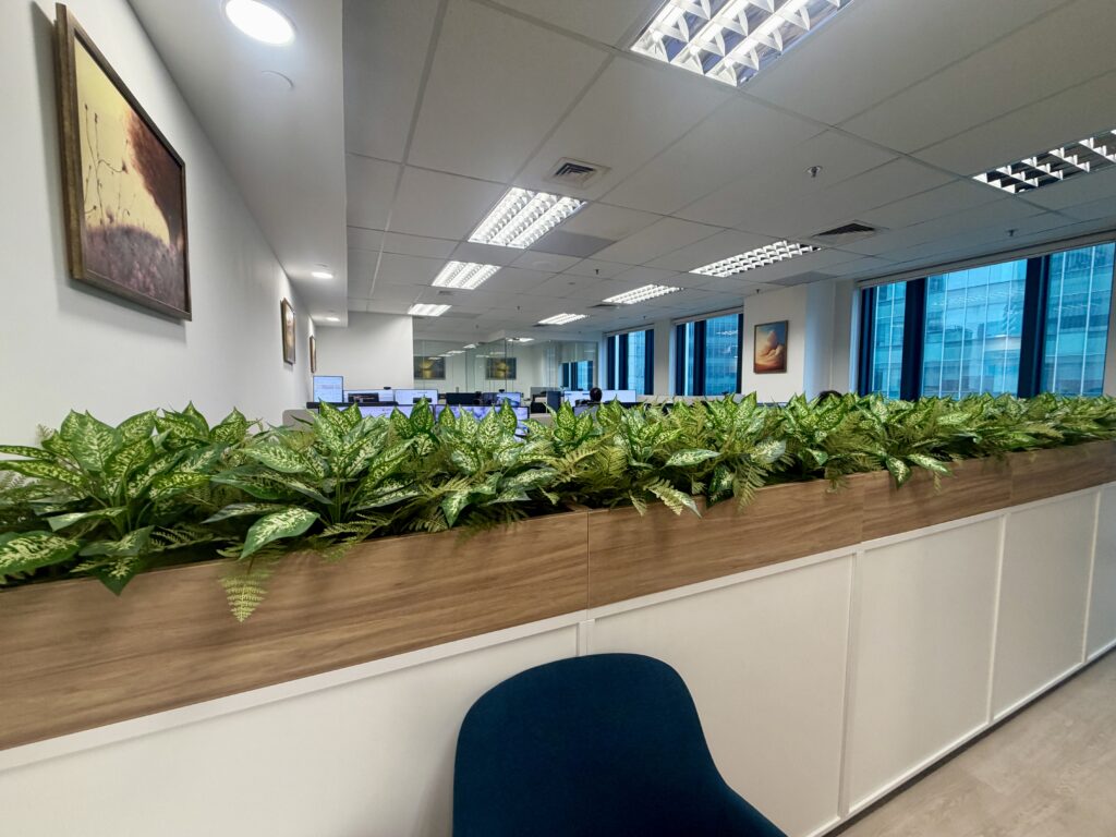 A row of potted green plants serves as a divider in an office, with workstations and windows visible in the background.