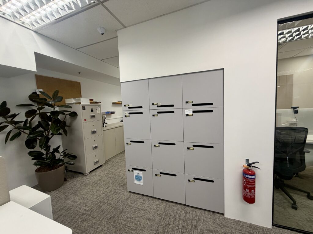 A wall of grey office storage lockers, a fire extinguisher mounted on the side, a potted plant, and a metal filing cabinet in an office space.