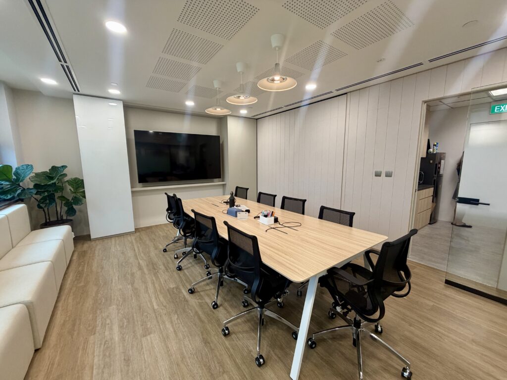 A conference room with a long wood-grain table, black swivel chairs, a wall-mounted television, and pendant lighting.