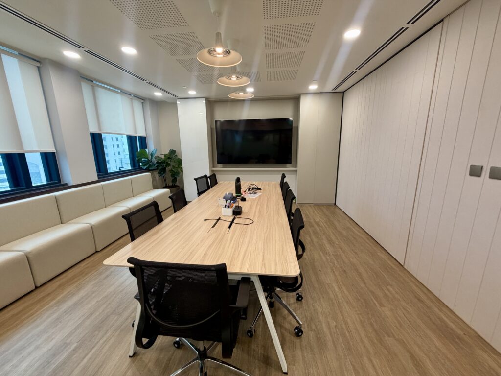 A conference room with a long wooden table, black mesh chairs, a wall-mounted flat-screen TV, and built-in bench seating along the window.