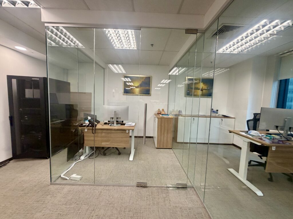 An office interior featuring two glass-walled cubicles, each equipped with a wooden desk, computer, and chair.